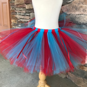 Puede incluir: Una falda tut&uacute; roja, azul claro y roja exhibida en un maniqu&iacute;. La falda est&aacute; hecha de capas de tul en colores alternos, con cintas rojas colgando. El tut&uacute; est&aacute; dise&ntilde;ado para ni&ntilde;os.