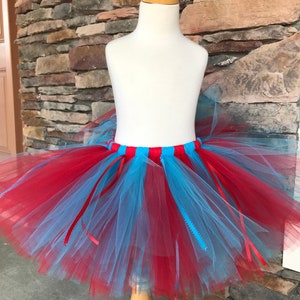 Puede incluir: Una falda tut&uacute; de tul rojo y azul claro sobre un maniqu&iacute;. La falda tiene una cintura roja y azul claro con cintas rojas. El tut&uacute; est&aacute; hecho de capas de tul rojo y azul claro.