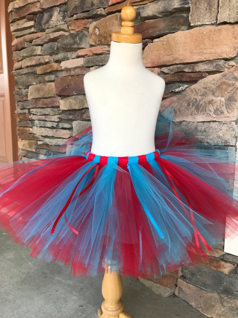 Puede incluir: Una falda tut&uacute; de tul rojo y azul claro sobre un maniqu&iacute;. La falda tiene una cintura roja y azul claro con cintas rojas. El tut&uacute; est&aacute; hecho de capas de tul rojo y azul claro.