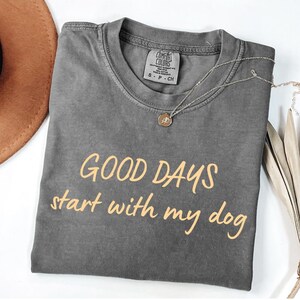 May include: A grey Comfort Colors t-shirt with the text "GOOD DAYS start with my dog" in light orange. A silver necklace with a small pendant is draped over the shirt.