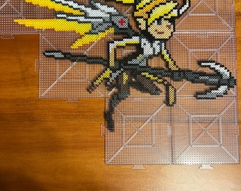 May include: A pixel art design of a video game character with yellow hair and wings, created with small plastic beads on a pegboard.