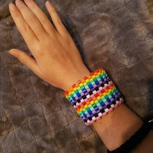 May include: A rainbow-coloured beaded bracelet with white and black beads. The bracelet is made of small, square-shaped beads.