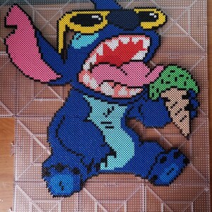 Puede incluir: Representación en pixel art de Stitch de Lilo & Stitch, hecha con cuentas azules, rosas, rojas, blancas y verdes. Stitch lleva gafas de sol amarillas y sostiene un cono de helado verde. La obra de arte está creada sobre una placa transparente.