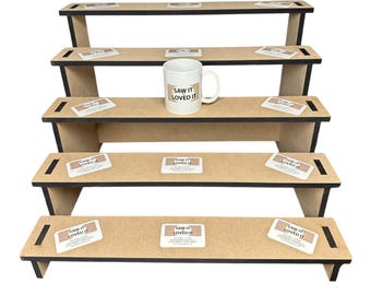 May include: A tiered display stand with five levels, constructed from light brown wood with black edges. A white mug with the text "SAW IT LOVED IT" sits on one shelf. Small rectangular stickers with the same text are placed on each level.