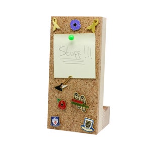 Puede incluir: Un tablero de corcho con un marco de madera y un soporte. El tablero de corcho tiene una nota adhesiva amarilla con la palabra "Stuff!" escrita en ella. Hay varios pines y insignias en el tablero de corcho, incluyendo una amapola morada, una cinta amarilla, una p&uacute;a de guitarra y una amapola roja.