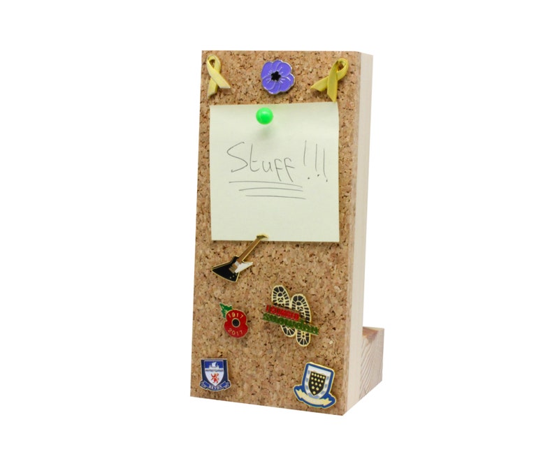 Puede incluir: Un tablero de corcho con un marco de madera y un soporte. El tablero de corcho tiene una nota adhesiva amarilla con la palabra "Stuff!" escrita en ella. Hay varios pines y insignias en el tablero de corcho, incluyendo una amapola morada, una cinta amarilla, una p&uacute;a de guitarra y una amapola roja.