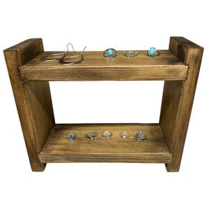 May include: A two-tiered, rustic wooden display stand with a dark stain. The top shelf holds silver earrings and rings with turquoise stones. The bottom shelf displays more rings with turquoise and other designs. The stand is ideal for showcasing jewelry.