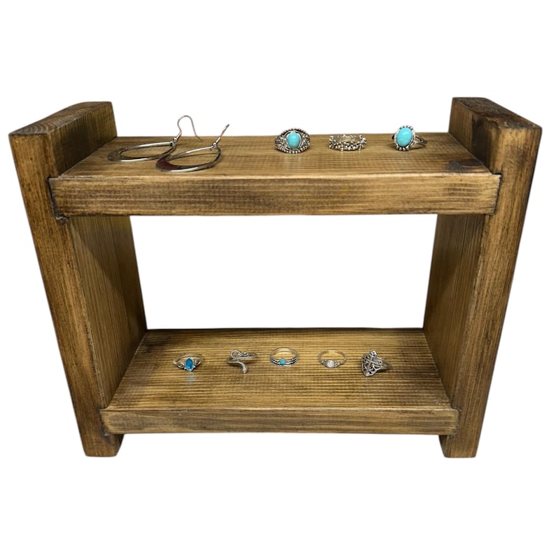 May include: A two-tiered, rustic wooden display stand with a dark stain. The top shelf holds silver earrings and rings with turquoise stones. The bottom shelf displays more rings with turquoise and other designs. The stand is ideal for showcasing jewelry.