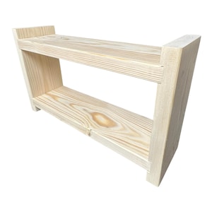 May include: A two-tiered, unfinished wooden shelf. The shelf is constructed from light-colored wood, featuring visible grain patterns. The design includes two horizontal shelves supported by angled side pieces, creating a simple, functional structure.