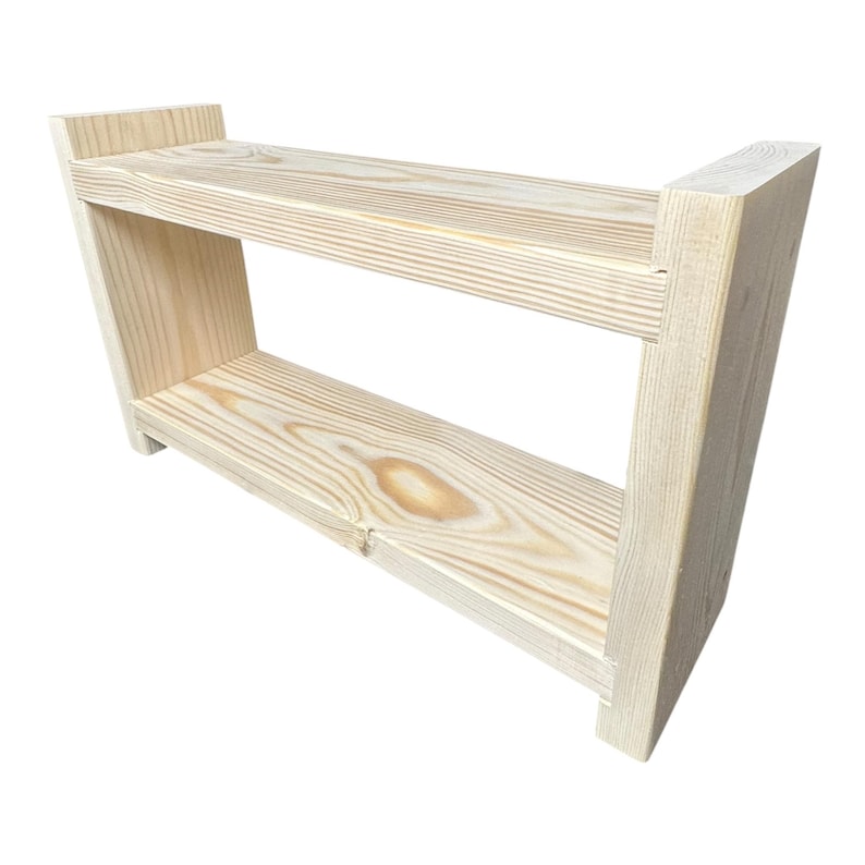 May include: A two-tiered, unfinished wooden shelf. The shelf is constructed from light-colored wood, featuring visible grain patterns. The design includes two horizontal shelves supported by angled side pieces, creating a simple, functional structure.