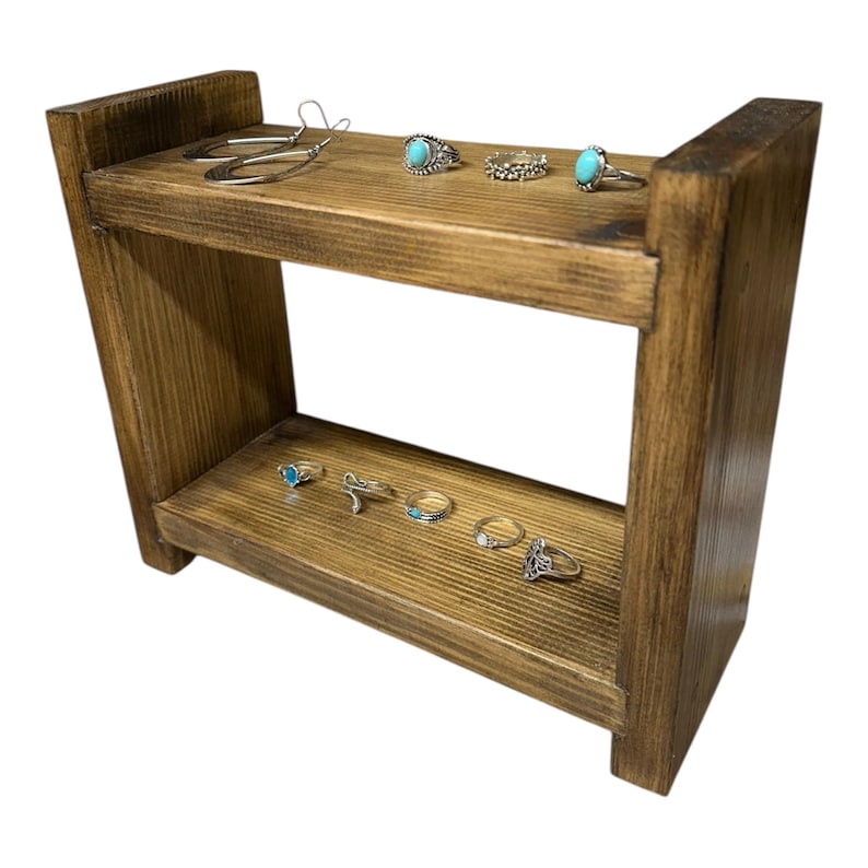 May include: A two-tiered, rustic wooden jewelry display stand. The stand is stained a warm brown and holds various rings and earrings. The rings feature turquoise stones and intricate designs. The earrings are silver-toned hoops.