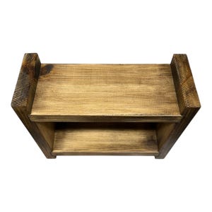 May include: A two-tiered, rectangular wooden shelf with a natural finish. The shelf has a simple design with two levels for storage or display. The wood grain is visible, and the edges are slightly darker, adding depth to the piece.