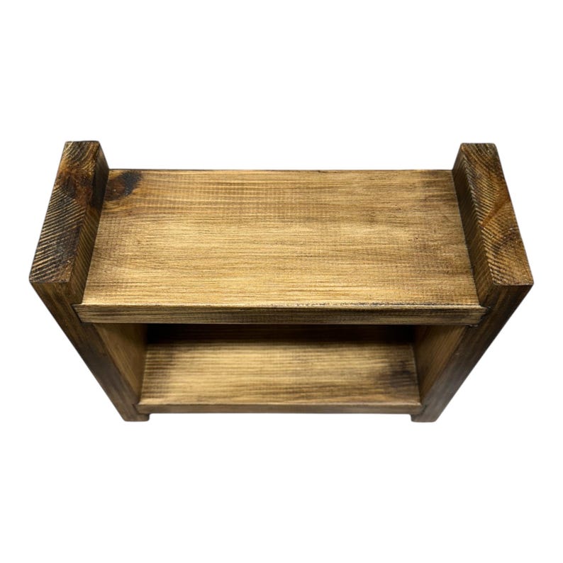 May include: A two-tiered, rectangular wooden shelf with a natural finish. The shelf has a simple design with two levels for storage or display. The wood grain is visible, and the edges are slightly darker, adding depth to the piece.