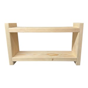 May include: A light-colored, two-tiered wooden shelf with a rectangular design. The shelf has a natural wood grain finish and is constructed with visible joints. The sides are angled, and the shelf is set against a white background.