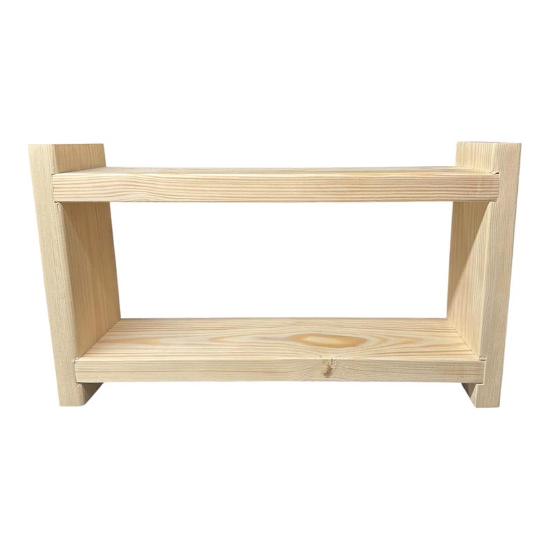 May include: A light-colored, two-tiered wooden shelf with a rectangular design. The shelf has a natural wood grain finish and is constructed with visible joints. The sides are angled, and the shelf is set against a white background.
