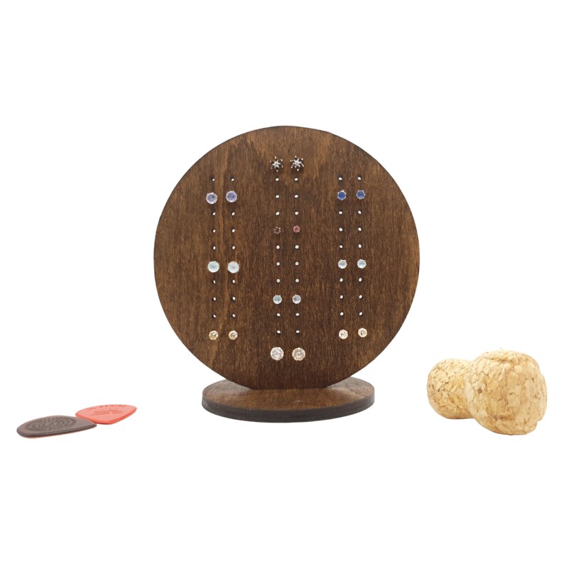 May include: A wooden earring display stand with a circular base and a round top with multiple holes for holding earrings. The stand is brown and has a variety of colourful earrings displayed on it.