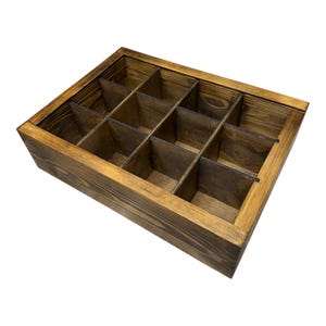 May include: A wooden storage box with a dark brown stain and a grid of twelve compartments. The box displays visible wood grain and has a rectangular shape. The compartments are evenly sized, suitable for organizing small items. The box is made of wood.