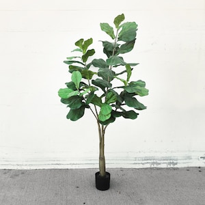 May include: An artificial fiddle leaf fig tree with large, vibrant green leaves. The plant has a brown trunk and is potted in a black container. The tree stands against a white wall and a grey concrete floor.