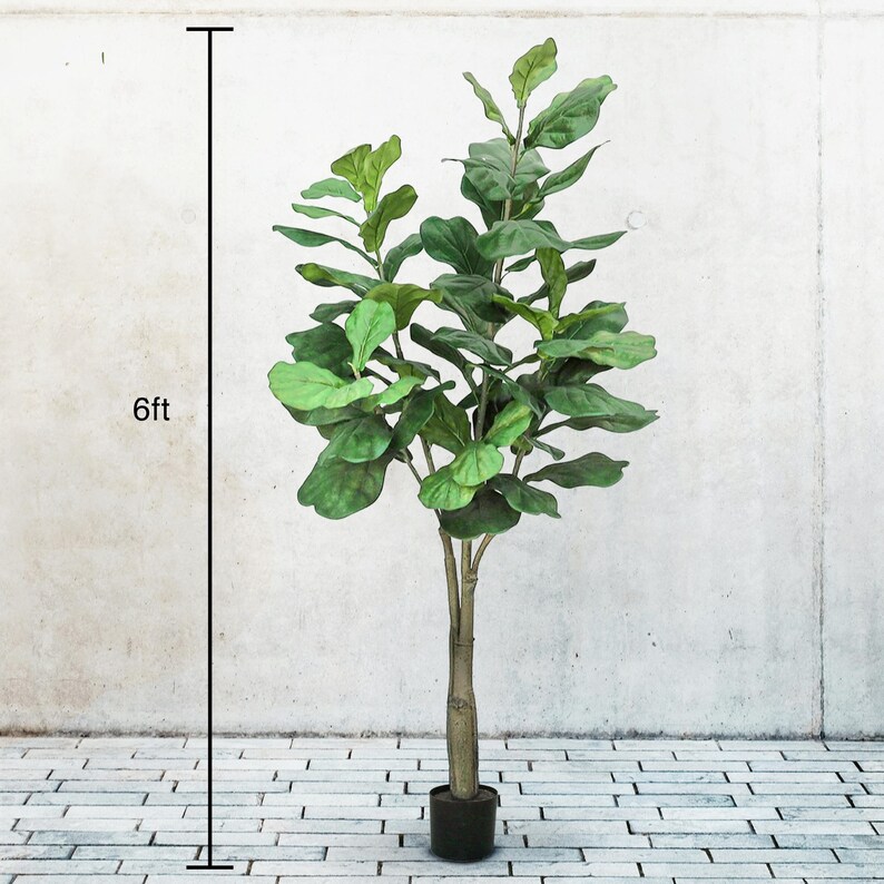 May include: An artificial Fiddle Leaf Fig tree, approximately 183cm tall, in a black pot. The tree features large, green leaves and a brown trunk. It stands against a white wall and a brick-patterned floor.