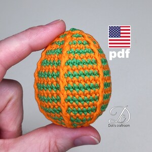 Può includere: Un ornamento a forma di uovo lavorato a uncinetto di colore arancione e verde. L'uovo è a strisce di filato arancione e verde e ha una superficie testurizzata. L'uovo è tenuto da una mano. L'immagine include anche una bandiera americana e la parola "pdf".
