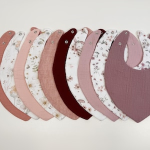 Puede incluir: Una colección de diez baberos para bebé en forma de media luna dispuestos en fila. Los baberos presentan una variedad de colores sólidos, incluyendo rosa empolvado, burdeos intenso y malva, junto a estampados florales sobre tela blanca.