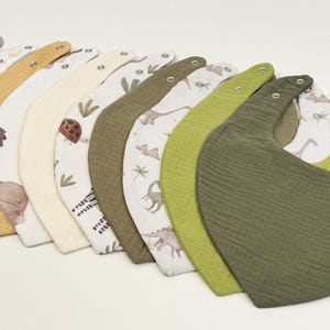 Puede incluir: Una colección de diez baberos para bebé en forma de media luna dispuestos en fila. Los baberos presentan una variedad de colores sólidos que incluyen verde oliva, amarillo mostaza y beige, junto a baberos blancos con estampados de animales.