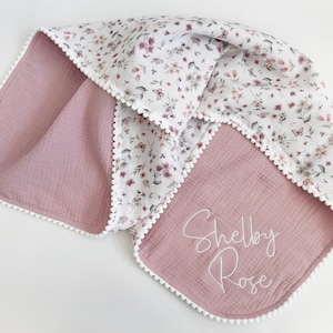 May include: A baby blanket with a floral pattern on a white background, and a solid mauve color on the reverse side. The blanket has a white pom-pom trim and the name "Shelby Rose" is embroidered on the mauve side.