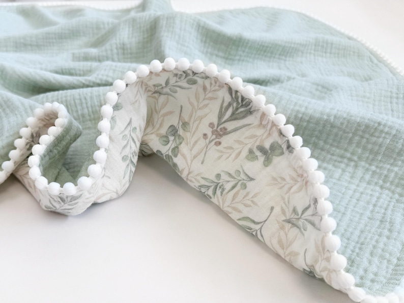 May include: A light green muslin blanket with a white pom-pom trim. The blanket's interior features a botanical print with green and brown leaves on a white background. The blanket is folded, revealing the patterned lining.