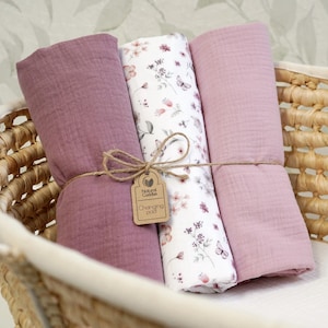 May include: Three rolled baby changing pads in a wicker basket. Two pads are solid mauve, and one is white with a floral and butterfly pattern. A tag with the words "Natural Cuddles Changing pad" is tied with twine.