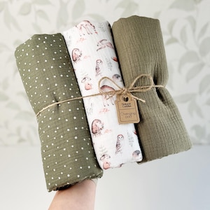 Könnte beinhalten: Drei gerollte Musselintücher, zusammengebunden mit Bindfaden und einem Pappanhänger mit der Aufschrift Natural Cuddles Changing Pad. Ein Tuch ist olivgrün mit weißen Punkten, eines ist weiß mit Waldtiermotiv und eines ist einfarbig olivgrün.