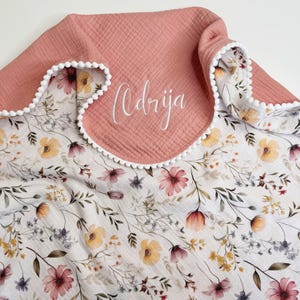 May include: A soft muslin blanket featuring a white base with a delicate floral pattern in shades of pink, yellow, and green. The blanket has a solid dusty rose fabric section at the top edge, accented with white pom-pom trim and the name Odrija.