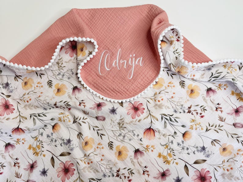 May include: A soft muslin blanket featuring a white base with a delicate floral pattern in shades of pink, yellow, and green. The blanket has a solid dusty rose fabric section at the top edge, accented with white pom-pom trim and the name Odrija.