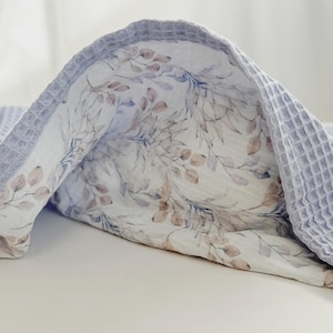 May include: A dual-textured blanket featuring a light blue waffle-knit border and a central panel with a botanical print. The fabric displays a pattern of soft brown and muted blue leaves and stems on a white background, creating a layered look.