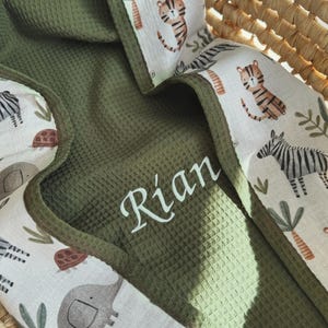 Puede incluir: Manta de bebé verde oliva con textura de gofre, con el nombre "Rian" impreso en blanco. La manta tiene un borde estampado con cebras, tigres, elefantes y follaje. La manta está en una cesta tejida.
