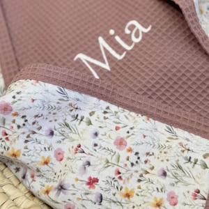 May include: A dusty rose waffle-textured fabric blanket featuring the name Mia embroidered in white script. The blanket is trimmed with a white fabric border decorated with a colorful wildflower print consisting of small pink, yellow, and purple flowers.