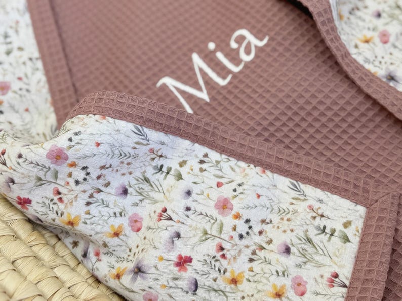 May include: A dusty rose waffle-textured fabric blanket featuring the name Mia embroidered in white script. The blanket is trimmed with a white fabric border decorated with a colorful wildflower print consisting of small pink, yellow, and purple flowers.