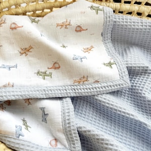 May include: A baby blanket featuring a white fabric side with a repeating pattern of small, colorful airplanes and helicopters in shades of green, orange, and blue, finished with a light blue waffle-textured fabric border and backing.
