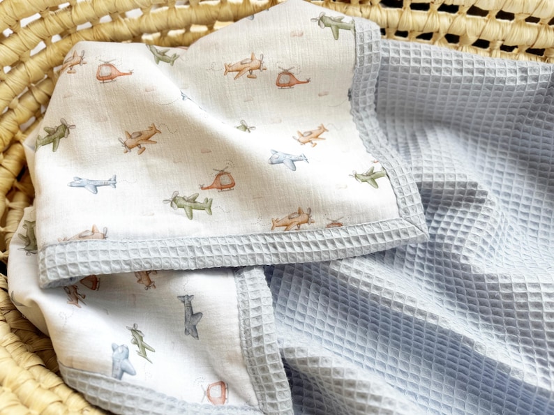 May include: A baby blanket featuring a white fabric side with a repeating pattern of small, colorful airplanes and helicopters in shades of green, orange, and blue, finished with a light blue waffle-textured fabric border and backing.