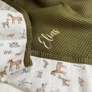 May include: A waffle-textured olive green blanket featuring a white cotton border with a farm-themed print of horses, geese, and tractors. The name Elias is embroidered in a light yellow cursive script on the olive green waffle fabric section.