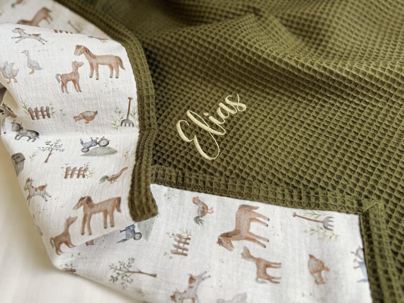May include: A waffle-textured olive green blanket featuring a white cotton border with a farm-themed print of horses, geese, and tractors. The name Elias is embroidered in a light yellow cursive script on the olive green waffle fabric section.