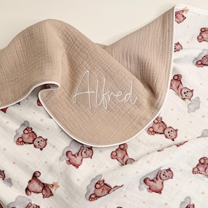 May include: A beige and white baby blanket with a teddy bear pattern. The blanket is reversible, with a solid beige side and a white side printed with brown teddy bears, clouds, and stars. The name "Alfred" is written in cursive on the beige side.