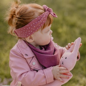 Puede incluir: Una ni&ntilde;a con una chaqueta rosa y una bufanda malva sostiene un conejito de peluche rosa. El conejito tiene orejas largas y una cara bordada sencilla. La ni&ntilde;a tambi&eacute;n lleva una diadema de lunares. El fondo es verde borroso.