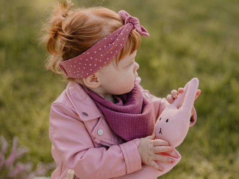 Puede incluir: Una ni&ntilde;a con una chaqueta rosa y una bufanda malva sostiene un conejito de peluche rosa. El conejito tiene orejas largas y una cara bordada sencilla. La ni&ntilde;a tambi&eacute;n lleva una diadema de lunares. El fondo es verde borroso.