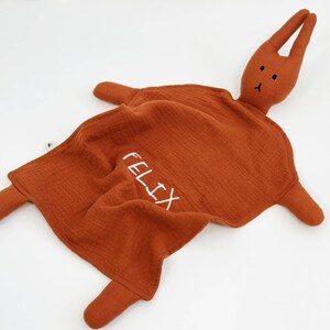 Puede incluir: Una mantita de seguridad para beb&eacute; de color &oacute;xido con una cabeza y extremidades de conejo de peluche. La palabra "FELIX" est&aacute; bordada en blanco en la manta. El conejo tiene peque&ntilde;os ojos negros y una nariz cosida.