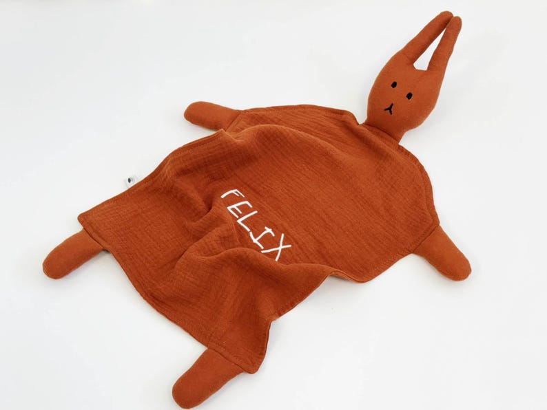Puede incluir: Una mantita de seguridad para beb&eacute; de color &oacute;xido con una cabeza y extremidades de conejo de peluche. La palabra "FELIX" est&aacute; bordada en blanco en la manta. El conejo tiene peque&ntilde;os ojos negros y una nariz cosida.