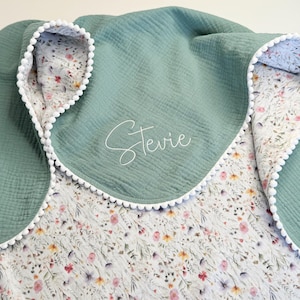 May include: A sage green baby blanket with a floral print interior and white pom-pom trim. The blanket features the name "Stevie" embroidered in a delicate script. The blanket is made of a soft, textured fabric.