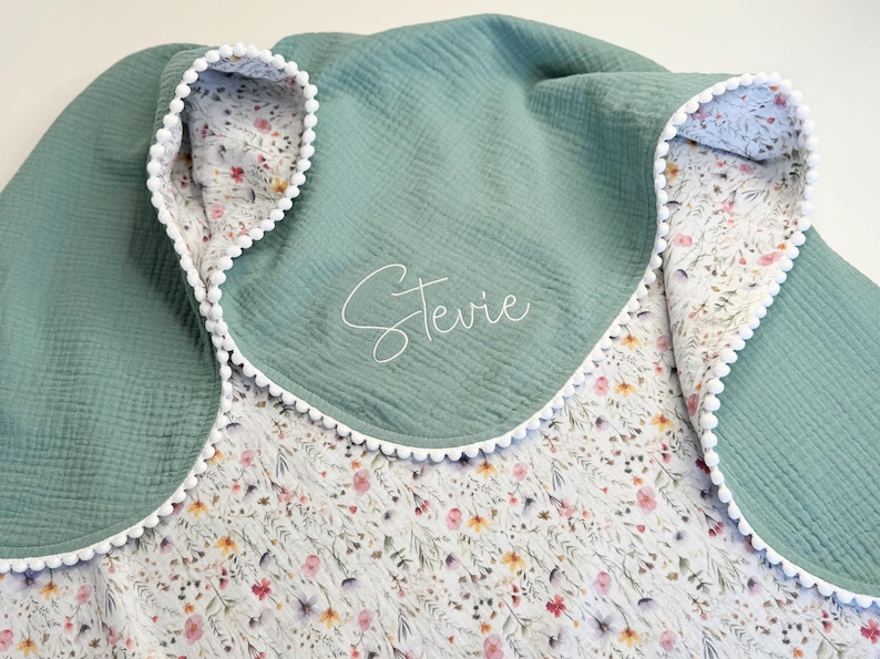May include: A sage green baby blanket with a floral print interior and white pom-pom trim. The blanket features the name "Stevie" embroidered in a delicate script. The blanket is made of a soft, textured fabric.