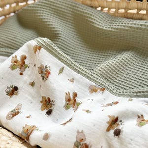 May include: A dual-textured blanket featuring a white fabric side with a repeating pattern of brown squirrels, acorns, and small green leaves, paired with a solid sage green waffle-knit fabric side, folded to display both distinct material surfaces.