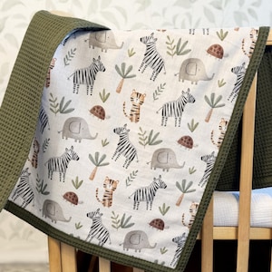 May include: A baby blanket featuring a white background with a repeating pattern of gray elephants, black and white striped zebras, orange and black tigers, brown turtles, and green palm trees, finished with a solid olive green waffle-textured border.