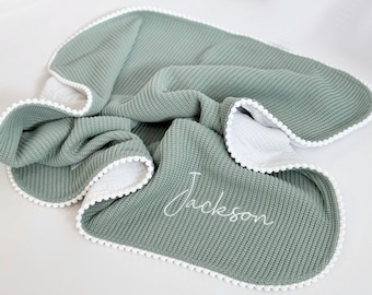 May include: A sage green waffle knit baby blanket with a white pom-pom trim and the name "Jackson" embroidered in white.